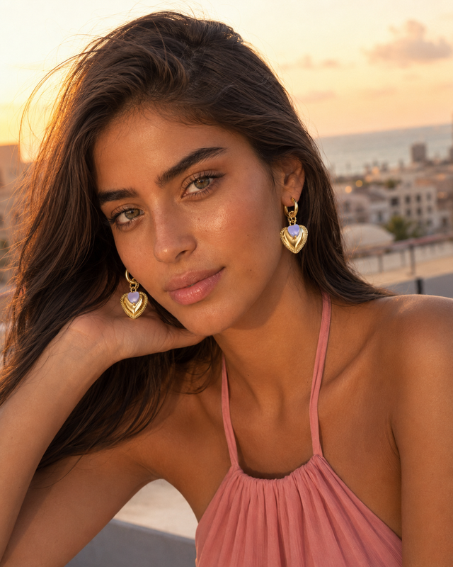 Femme méditerranéenne portant les boucles d’oreilles pendantes cœur doré et cristal lilas Lumé dans un décor côtier ensoleillé